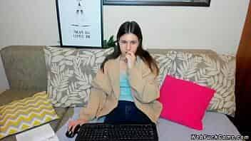 Brunette amateur teen sitting in clothes on the couch and typing on her keyboard while getting remote controlled vibrations from users of webcam show video